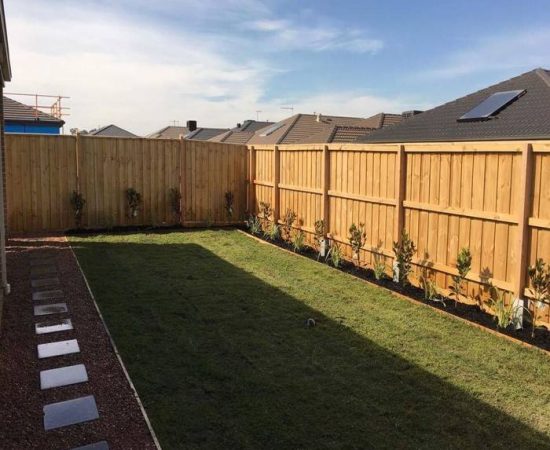 A backyard with a wooden fence, concrete pavers, natural turfing and a garden bed.